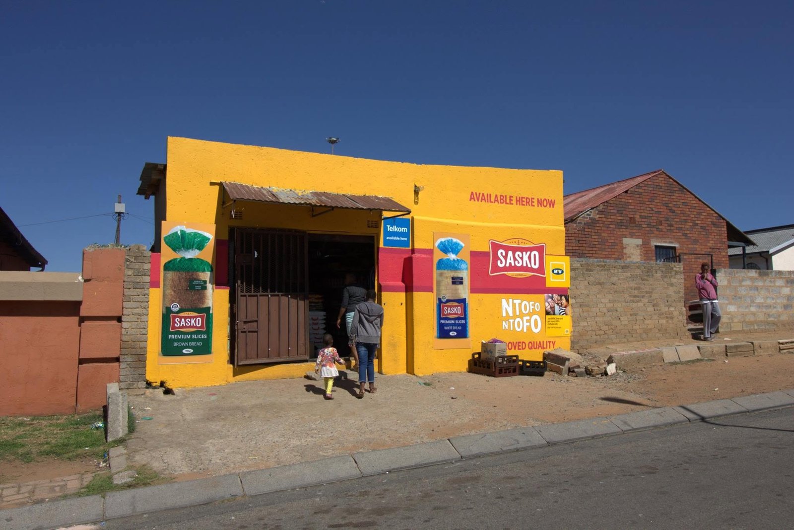 Township bezoeken in Zuid-Afrika: felgele spaza shop in Soweto met reclame op de gevel en buurtbewoners die naar binnen lopen